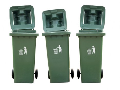 Three green wheeled wheelie bins with open lids arranged in a row on a paved surface, likely in an outdoor area adjacent to a property. The bins are made of textured plastic with white recycling symbols and a figure disposing of waste printed on their fronts. The background reveals a paved driveway or pathway, with the environment appearing clean and well-maintained, typical of residential or commercial waste collection settings in the Twickenham area. The bins are positioned side by side, with the middle bin slightly tilted, suggesting they are in use for rubbish or recycling collection services by Business Waste Removal Twickenham. The lighting indicates natural daylight, and the overall scene emphasizes a tidy presentation of waste management infrastructure suitable for local rubbish removal needs.