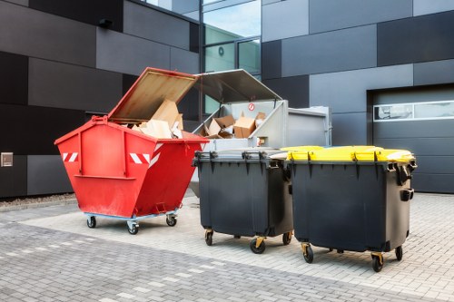 The image shows three large wheelie bins positioned on a paved outdoor area adjacent to a modern building with dark grey and black cladding. The first bin on the left is red with a partially open lid, revealing an interior filled with assorted cardboard boxes and packaging waste. The middle bin is dark grey with a yellow-lidded lid, closed and situated next to the red bin. The third bin, also dark grey with a closed lid, is placed slightly behind the others. The ground surface is made of grey paving stones, and the background features the building's reflective glass windows and sleek metal panels. The scene is clear and well-lit, emphasizing the functional nature of the waste containers, which are typically used for rubbish and waste collection services, such as those offered by Business Waste Removal Twickenham, servicing areas within Twickenham and the surrounding postcode regions.