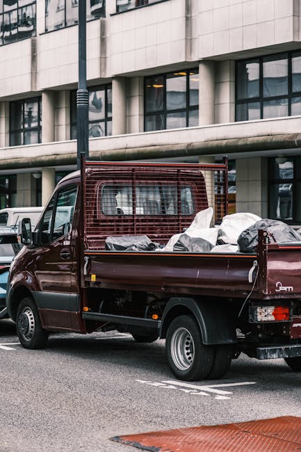 A small, dark red pickup truck parked on a city street in front of a modern building with large windows and a concrete facade. The truck's open bed is loaded with various types of waste and rubbish, i