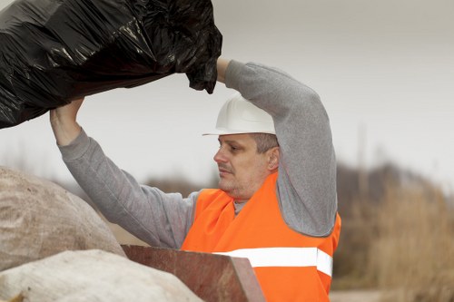 A male waste collection worker wearing a white safety helmet and an orange high-visibility safety vest with reflective stripes is standing outdoors in a rural or industrial area, surrounded by a collection of large black rubbish bags and various discarded materials. The worker is lifting a black rubbish bag above his head, preparing to deposit it into a waste collection vehicle or designated disposal point. In the background, there are blurred natural elements and overcast sky, indicating daytime with neutral weather conditions. The scene is typical of rubbish removal operations in Twickenham, with the worker demonstrating professional waste handling procedures with textured black bags and sturdy work attire. The setting suggests an organized waste clearance activity, possibly part of a service provided by Business Waste Removal Twickenham, focusing on efficient rubbish collection and disposal in the local area.